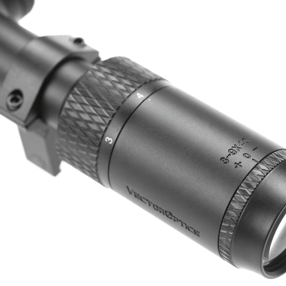 A close-up shot of a black Vector Optics rifle scope, showing the textured adjustment rings and the clear objective lens on a white background.